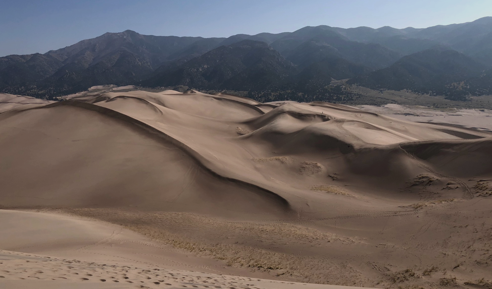 Expansive sand dunes with footprints leading towards distant mountains under a clear blue sky.