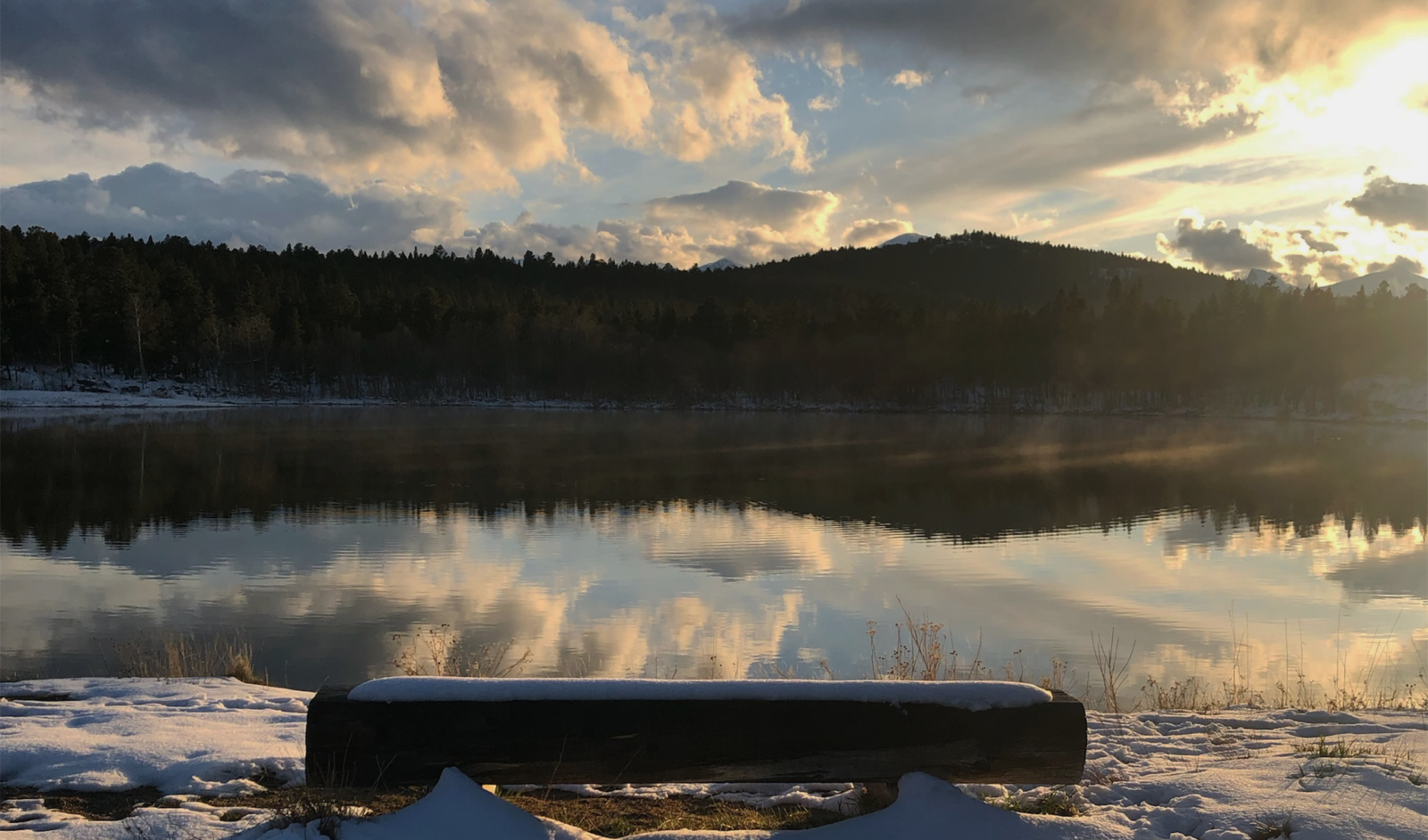 Snow-covered shoreline with a wooden bench overlooking a calm lake reflecting the sunset sky and surrounding forested hills.