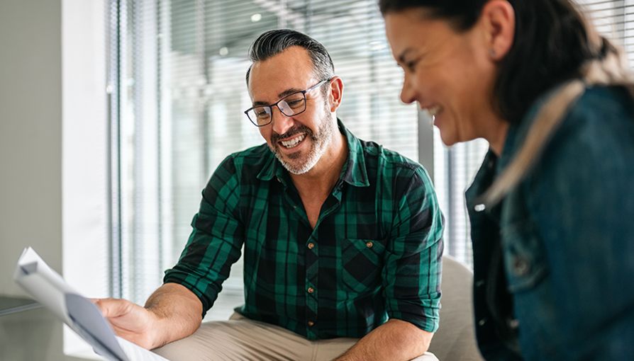 Smiling financial advisor in glasses and a green plaid shirt discusses paperwork with a client in a modern office setting.