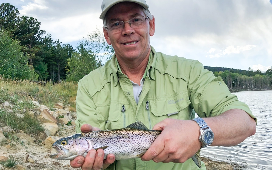 Man wearing a green shirt and cap proudly holding a freshly caught trout by a lakeshore, surrounded by trees.